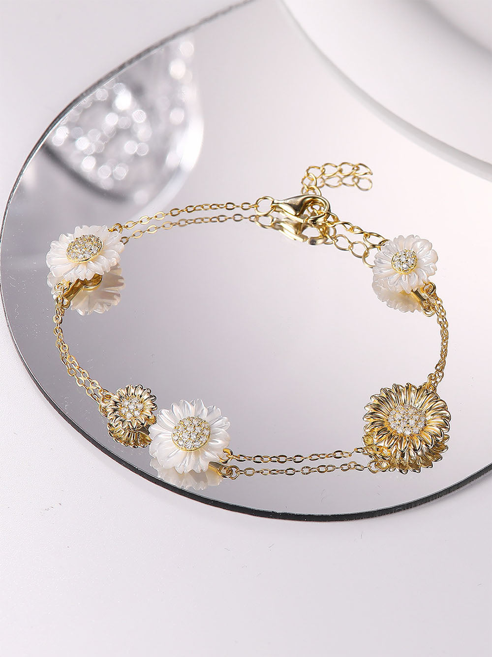 The Mother of Pearl Double Sun Flower Bracelet features 5A cubic zirconia stones and white Mother of Pearl in a delicate flower design. The bracelet has a 6-inch chain, with a 1-inch (2 cm) charm, and is secured with a lobster clasp. It is from the brand Joy & Rachel.